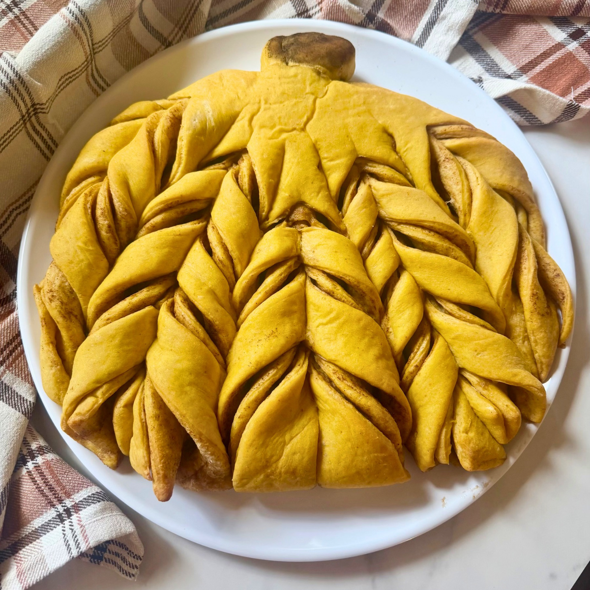 A large sourdough braided pumpkin pie or pumpkin bread with a decorative top, served on a white plate with a plaid napkin in the background.